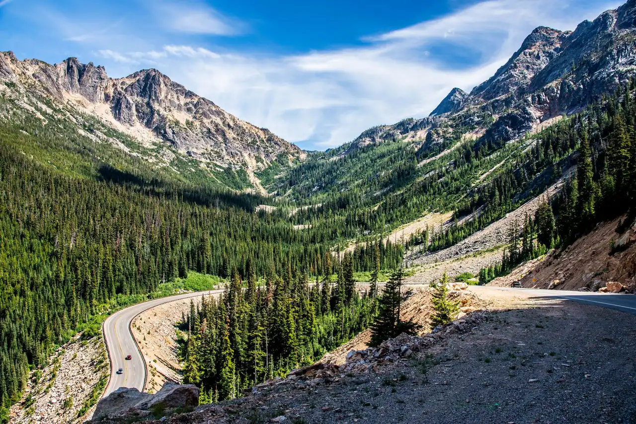 Two-lane mountain highway descending through dense conifer forest toward jagged snow-streaked peaks