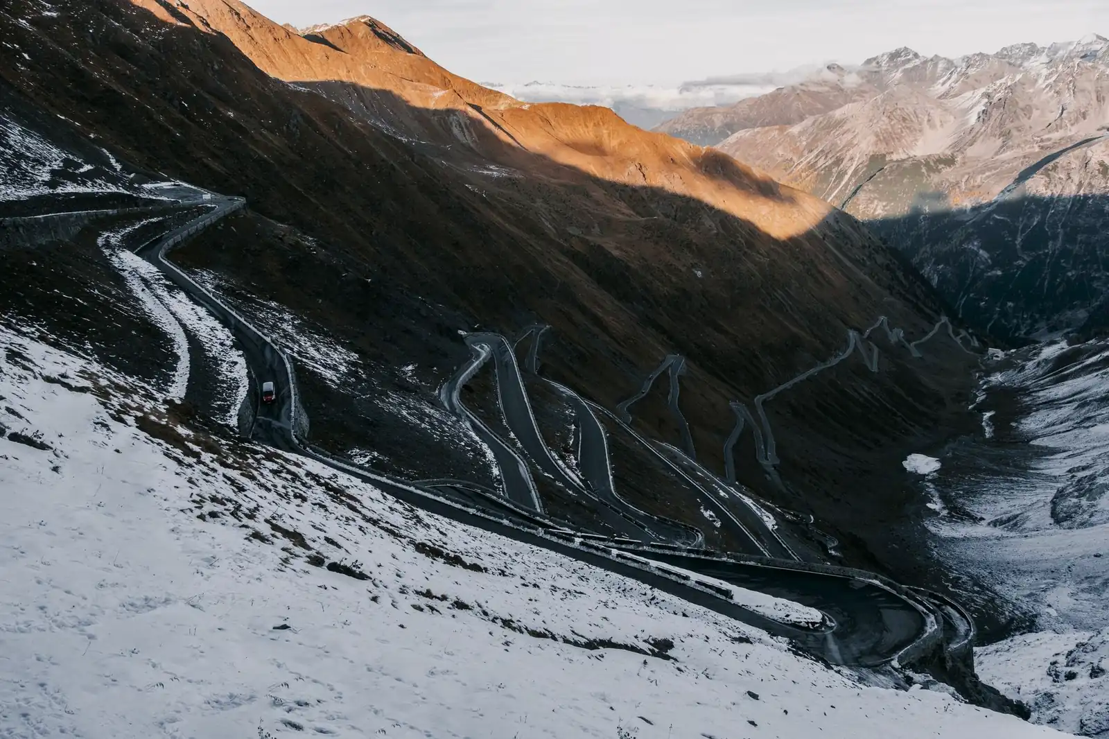 Aerial view of a mountain highway's hairpin switchbacks climbing a snow-dusted alpine headwall at golden hour
