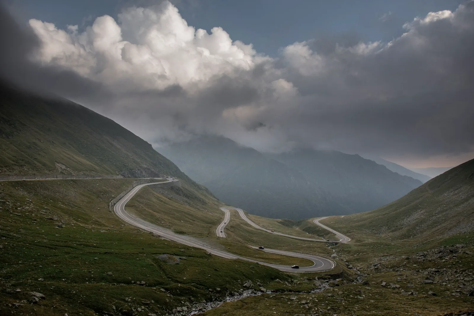 Dark storm clouds building over a winding mountain pass road with cars descending the switchbacks below
