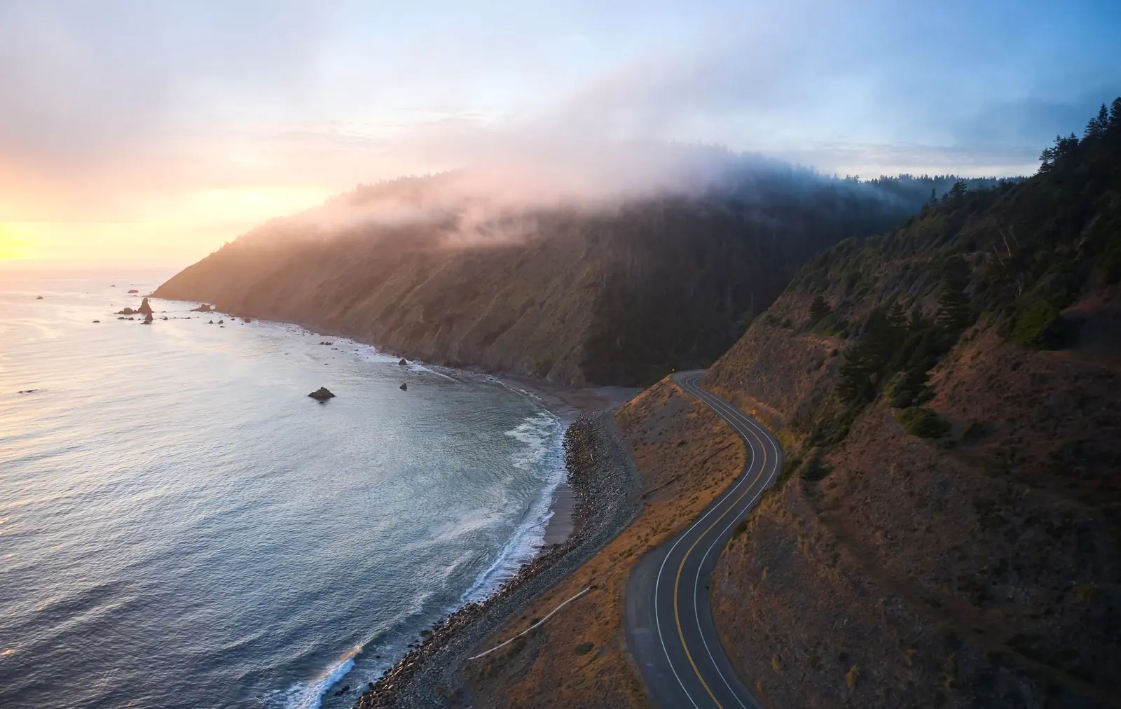 A two-lane highway hugging a misty, forested cliffside above the ocean at golden hour