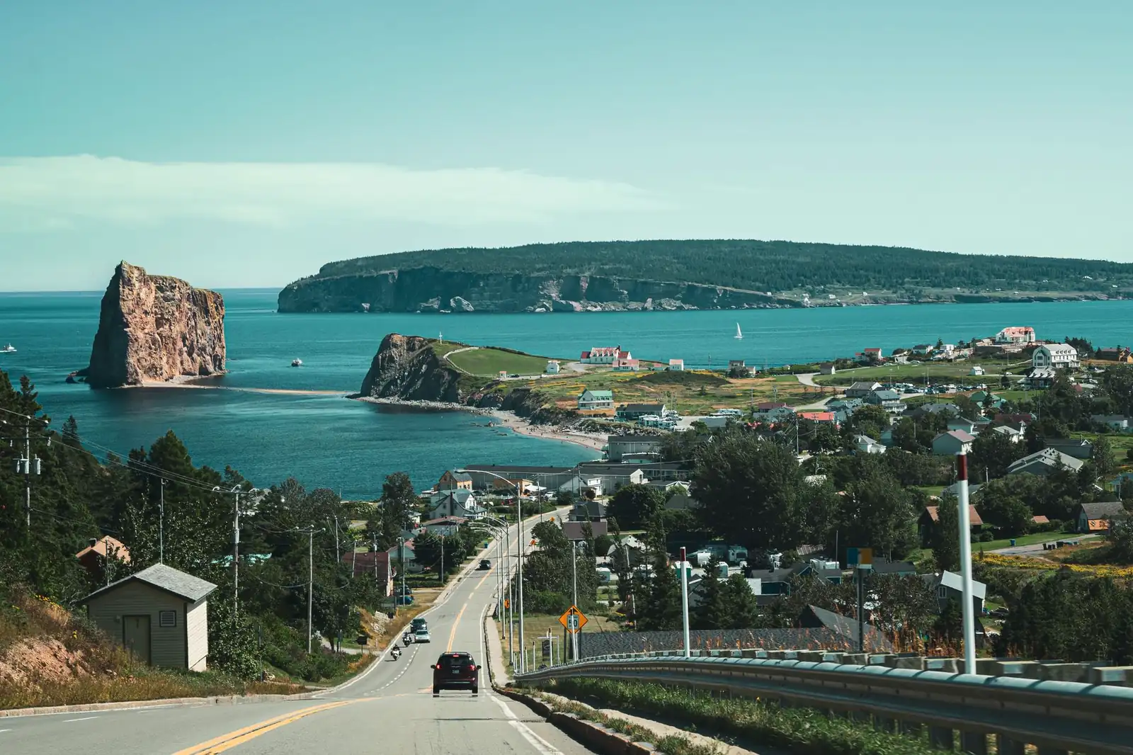 Route 132 winding down into Percé, Quebec, with Percé Rock and its sea arch visible in the bay beyond the village