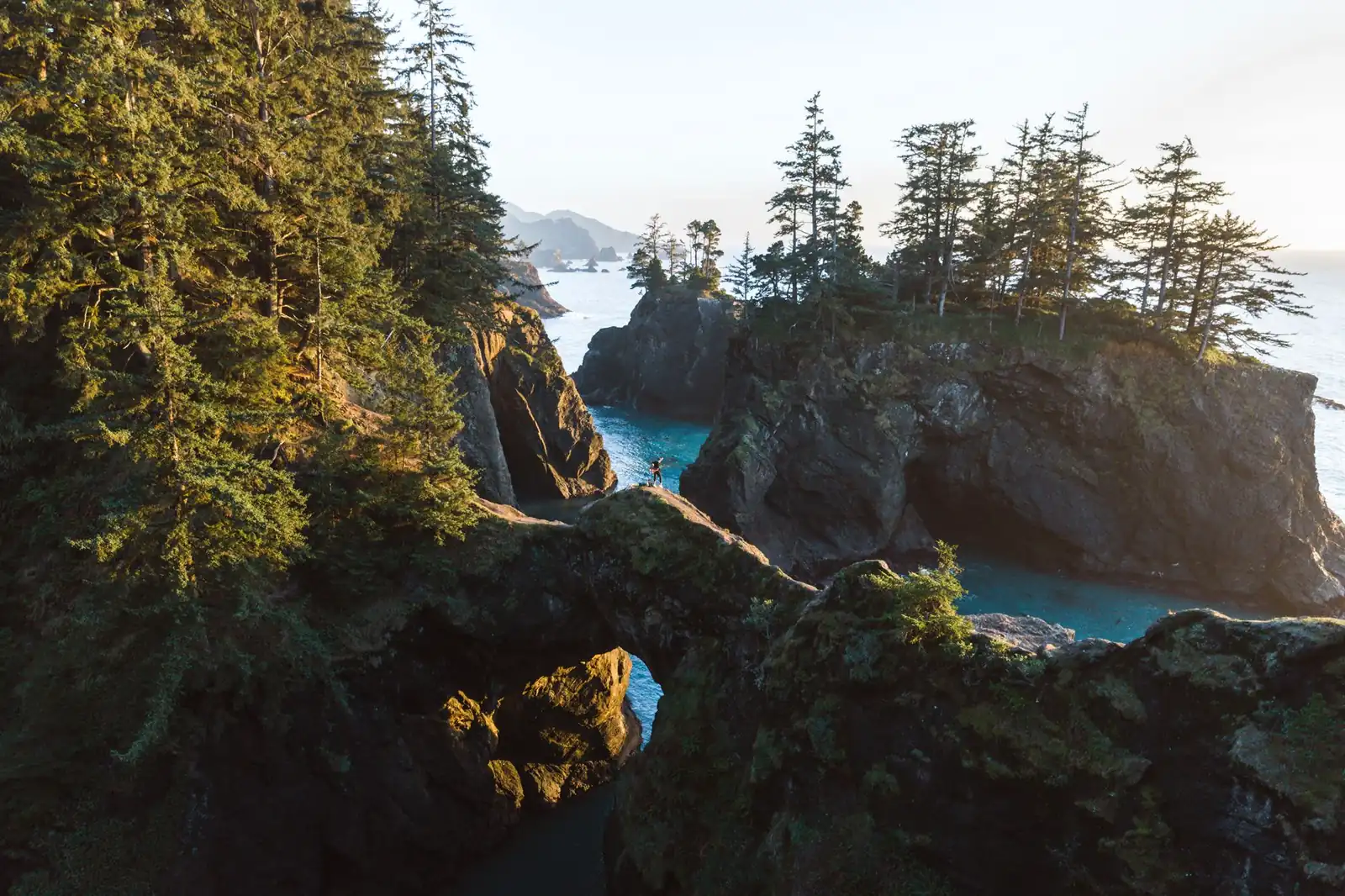 A hiker standing on the Natural Bridges rock arch above turquoise coves in the Samuel H. Boardman State Scenic Corridor