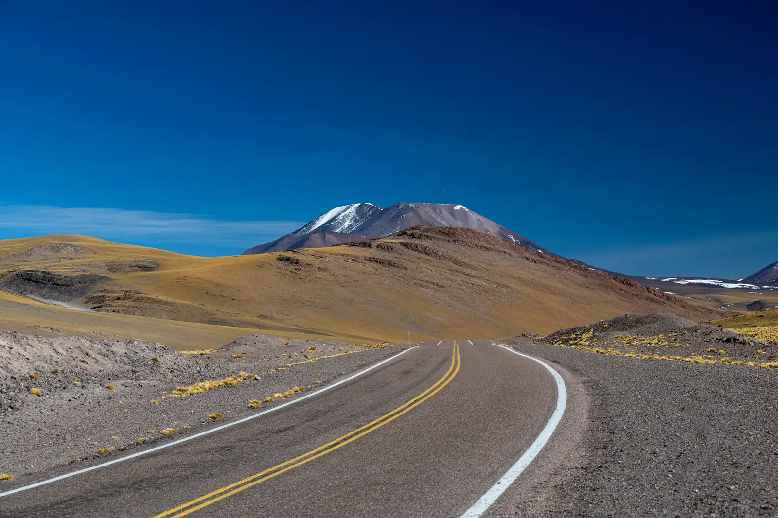 Paved two-lane highway running straight across a treeless high-alpine plateau toward a snow-capped peak, no guardrails