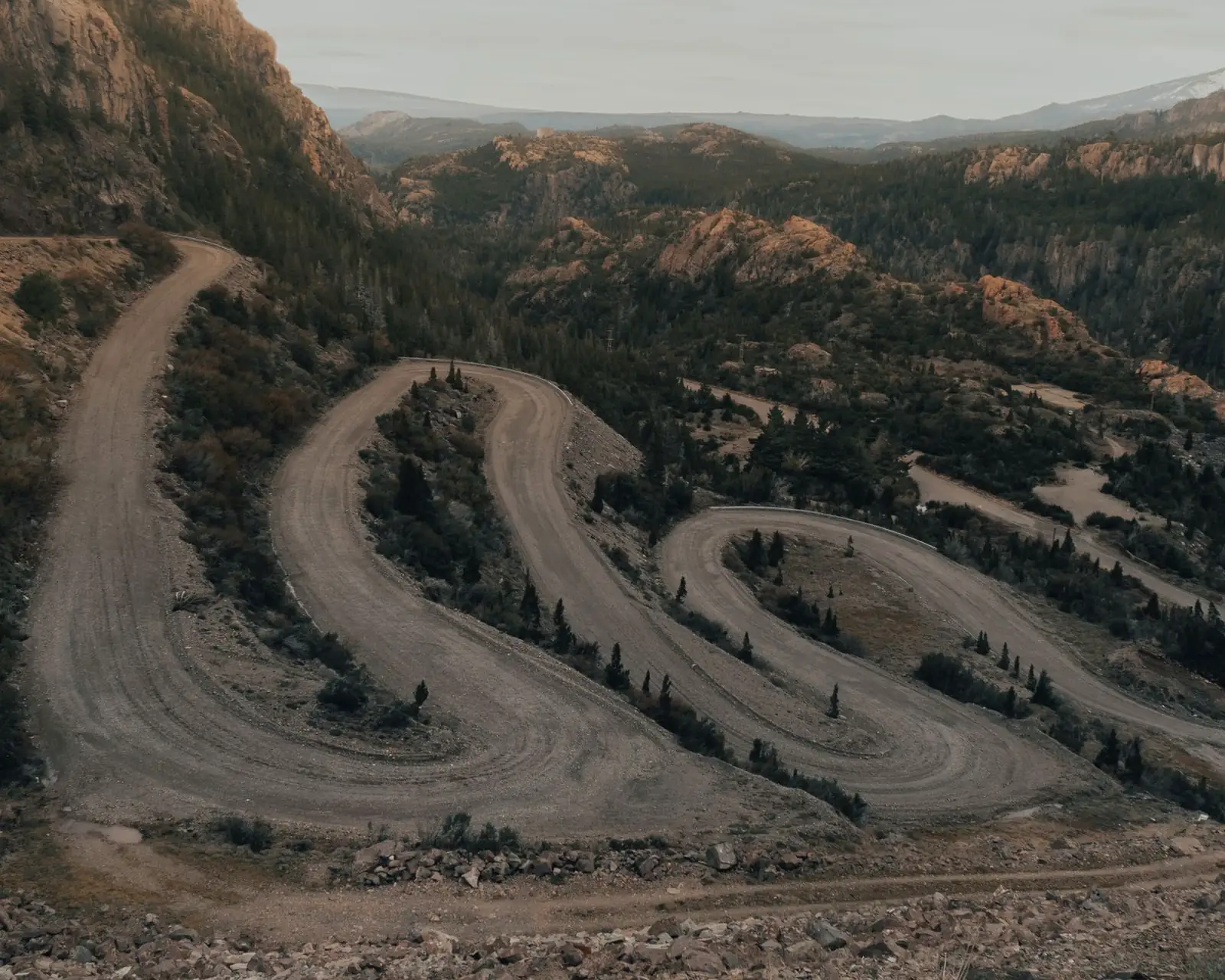 Aerial view of a narrow, unpaved switchback road climbing a forested mountain headwall in evening light
