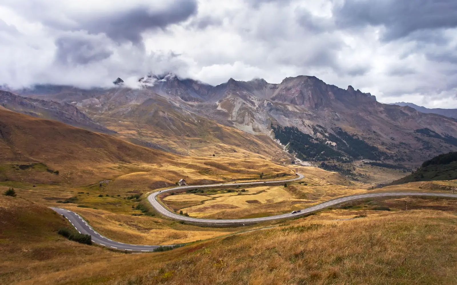 Paved mountain highway winding across a golden, treeless alpine valley with storm clouds building over the peaks ahead