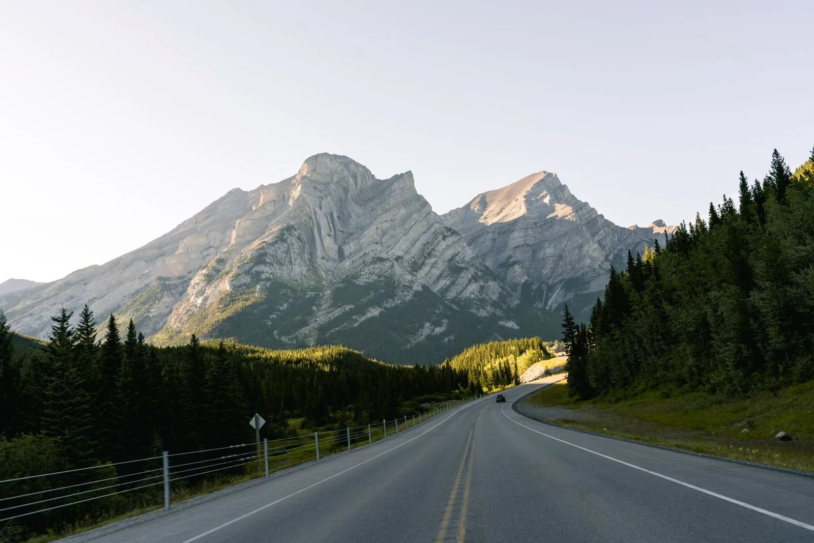 Highway 1 curving through the Canadian Rockies near Banff, evening light on layered rock peaks with a small car on the road for scale