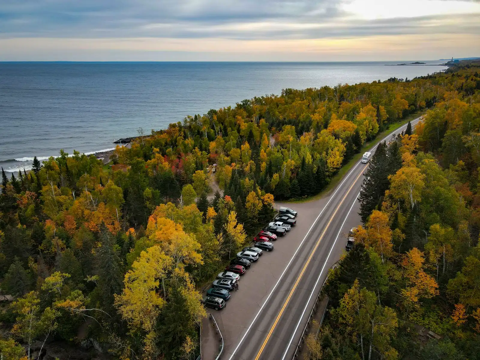 Aerial view of a two-lane highway following the Lake Superior shoreline through fall-colored forest, with a few cars and an RV on the road